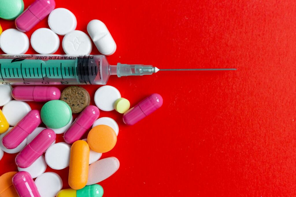 Top view of various colorful pills and a syringe on a vivid red surface, highlighting medical themes.